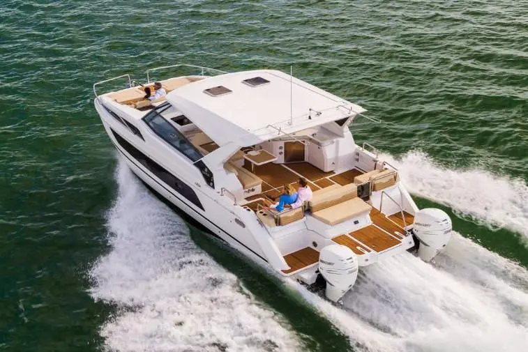 A luxury motor yacht speeds through green waters, with two people relaxing on deck under clear skies.